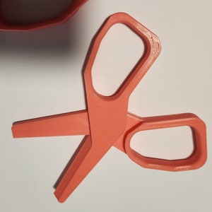 May include: A pair of bright orange plastic scissors with rounded edges. The scissors are open and ready to cut.