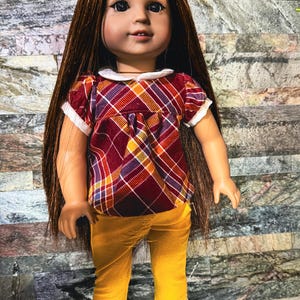 Könnte beinhalten: Eine Puppe mit langem, glattem braunem Haar, braunen Augen und heller Haut. Die Puppe trägt eine kurzärmelige Bluse mit einem bordeauxroten, gelben und weißen Karomuster und einem weißen Kragen, dazu eine senfgelbe Hose und weiße Schuhe.