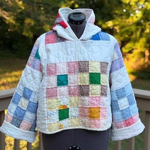 May include: A hooded pullover constructed from repurposed patchwork quilt fabric featuring a grid of multi-colored squares in shades of pink, yellow, green, blue, and white. The garment has long sleeves with wide cuffs and a quilted texture throughout.
