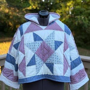 May include: A hooded pullover jacket constructed from quilted fabric featuring a patchwork star pattern. The design incorporates red and white checkered fabric, blue denim-style material, and white fabric with small blue floral prints.