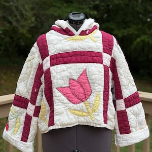 May include: A quilted hooded pullover made from off-white fabric with a grid pattern of deep magenta strips. The garment features appliquéd floral motifs in shades of pink and pale yellow, long sleeves, and a hood, all with visible stitching.