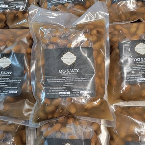 May include: Multiple clear plastic bags filled with boiled peanuts in a brine solution. Each bag has a black and white label that reads "OG Salty Boiled Peanuts" with instructions for heating and ingredients. The label also includes the net weight of 24oz (680.3g) and a batch number.