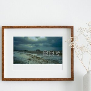 Könnte beinhalten: Ein gerahmtes Foto einer stürmischen Meereslandschaft mit einem Pier in der Ferne. Der Himmel ist dunkel und dramatisch, mit Wolken, die Schatten auf das Wasser werfen. Die Wellen schlagen an die Küste und erzeugen ein Gefühl von Bewegung und Energie.
