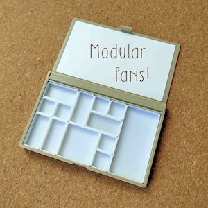 Pu&ograve; includere: Una tavolozza di plastica beige vuota con un coperchio bianco che dice "Modular Pans!" La tavolozza ha 12 pozzetti vuoti di diverse dimensioni.