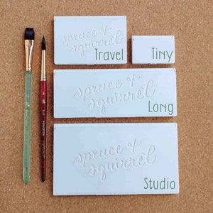 Pu&ograve; includere: Tre palette di acquerello bianche con il testo "spruce + squirrel" e le parole "Travel", "Long" e "Studio" scritte sotto. Due pennelli sono sul lato sinistro dell'immagine.