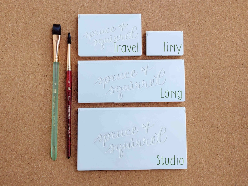 Pu&ograve; includere: Tre palette di acquerello bianche con il testo "spruce + squirrel" e le parole "Travel", "Long" e "Studio" scritte sotto. Due pennelli sono sul lato sinistro dell'immagine.