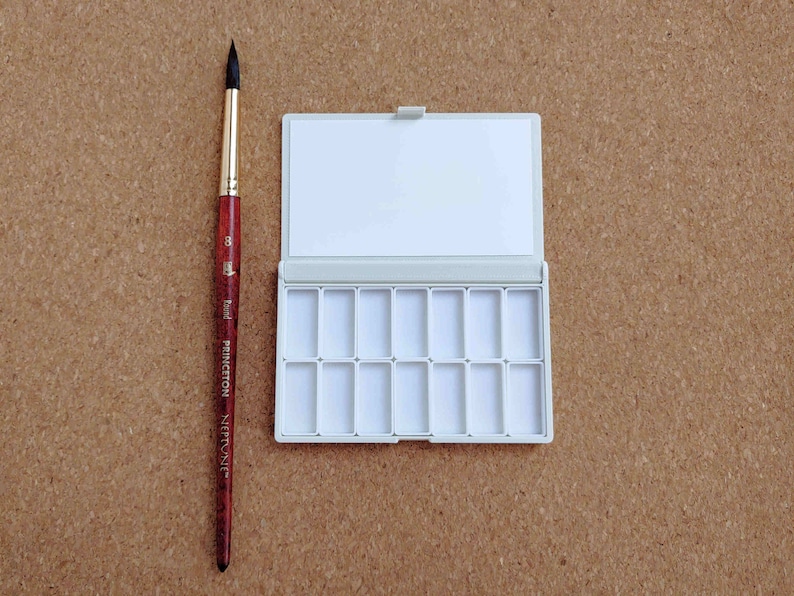 Pu&ograve; includere: Una tavolozza di colori ad acquerello bianca con 15 vaschette vuote, aperta e pronta per l'uso. Un pennello marrone e nero &egrave; posto accanto alla tavolozza.