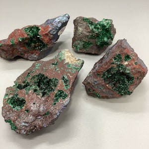 Puede incluir: Cuatro rocas ásperas, de color marrón oscuro, verde e iridiscente con cristales verdes. Las rocas están sobre una superficie blanca.