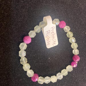 May include: A bracelet made of clear prehnite beads with pink ruby accents. The bracelet has a tag that reads "Prehnite Ruby".