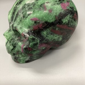 May include: A green and pink skull carved from a gemstone. The skull has a detailed face with teeth and a textured surface.