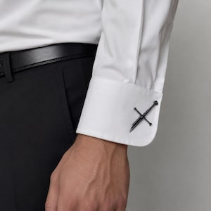 Op de afbeelding: Een wit overhemd met lange mouwen en een stijve manchet die is vastgezet met een donker metalen manchetknop in de vorm van een zwaard. Het overhemd zit in een zwarte broek die wordt vastgehouden door een zwarte leren riem. De manchetknop heeft een gedetailleerd ontwerp van een gevest en lemmet.