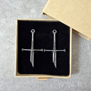 Könnte beinhalten: Ein Paar schwertförmige Anhänger aus silberfarbenem Metall mit Parierstange und einer Klinge, auf der vertikal der Schriftzug "STRENGTH" eingraviert ist. Die Anhänger liegen in einer quadratischen, mit schwarzem Samt ausgekleideten Schachtel.