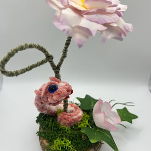 Peut inclure: Un serpent en cure-pipe rose avec un œil bleu et une langue rouge enroulé sur une base en tranche de bois circulaire recouverte de mousse verte. Une grande fleur rose pâle au centre jaune et une plus petite fleur rose avec des feuilles vertes.