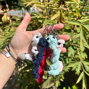 Op de afbeelding: Een set van vier spiraalvormige pluche sleutelhangers in grijs, donkerblauw, rood en lichtblauw, elk voorzien van een klein donker oog en een zilveren metalen ring. Het zachte, getextureerde garen geeft elk item een gedraaid, wormachtig uiterlijk.