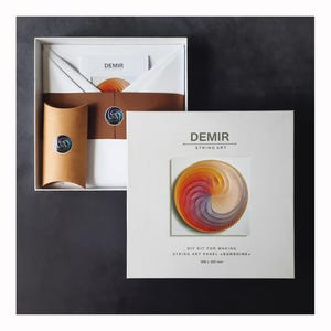 Könnte beinhalten: Ein weißes Box-Set mit einem DIY-String-Art-Kit. Die Box zeigt das "DEMIR"-Logo und ein fertiges String-Art-Panel in einem kreisförmigen Design mit Farbtönen von Orange, Gelb, Rosa und Lila. Das Kit enthält Anweisungen und Materialien für die Herstellung des 30 x 30 cm großen Panels.