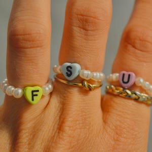 Könnte beinhalten: Drei Ringe an den Fingern. Ein Ring hat eine lindgrüne herzförmige Perle mit dem Buchstaben F, einer eine hellblaue Herzperle mit dem Buchstaben S und einer eine hellrosa Herzperle mit dem Buchstaben U. Alle Ringe enthalten weiße perlenartige Kügelchen.