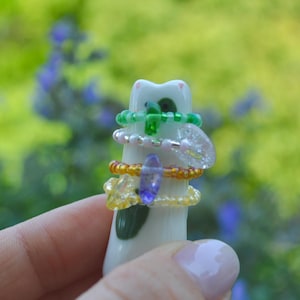 Könnte beinhalten: Eine weiße Keramikkatzenfigur mit grünen Flecken dient als Ständer für vier Perlenringe. Die Ringe bestehen aus verschiedenen Farben, darunter grüne, rosa, orange und gelbe Perlen, wobei einige kleine Anhänger und klare Akzente enthalten.