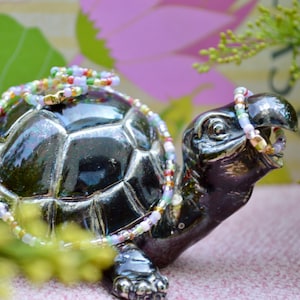 Könnte beinhalten: Eine glänzende schwarze Keramik-Schildkrötenfigur mit strukturiertem Panzer, geschmückt mit einer zarten Perlenkette aus kleinen, bunten Glasperlen in Rosa, Grün, Gelb und Weiß, die über den Rücken drapiert ist und im Maul gehalten wird.