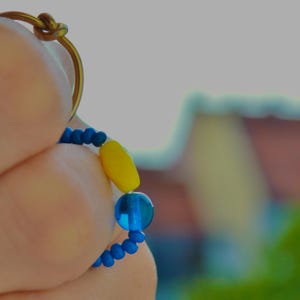 Pode incluir: Um anel com uma banda metálica fina e um detalhe de nó no topo. A banda é adornada com uma série de pequenas contas azuis, uma conta amarela maior e uma conta azul translúcida, criando um design circular colorido e texturizado.