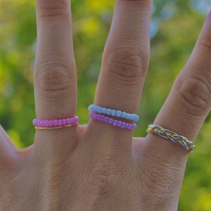 Può includere: Una collezione di anelli indossati su tre dita. Un dito presenta un anello di perline rosa sovrapposto a una sottile fascia dorata. Un altro dito mostra un anello di perline azzurre con uno viola. Un terzo dito indossa un anello in metallo intrecciato.