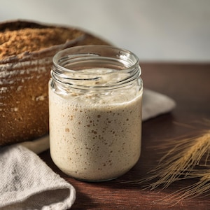 May include: A glass jar filled with a bubbly, light brown sourdough starter, next to a loaf of freshly baked sourdough bread and wheat stalks on a wooden surface.