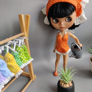 Puede incluir: Una muñeca con un vestido de lunares blancos y naranjas y una diadema a juego. La muñeca sostiene una pequeña regadera y está de pie junto a una planta suculenta en maceta. La muñeca también lleva zapatos naranjas.