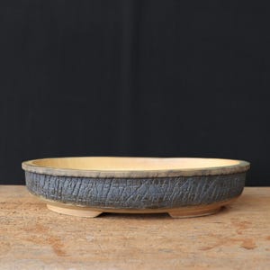 Op de afbeelding: Een handgemaakte, ovale bonsai pot. De buitenkant heeft een getextureerde, donkergrijze afwerking, terwijl de binnenkant een lichtgele tint heeft. De rand is een lichtere tint bruin. De pot staat op kleine, rechthoekige voetjes.