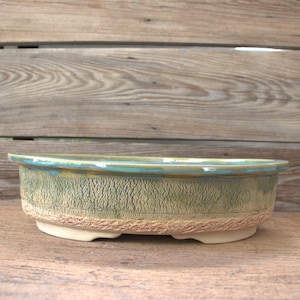 Könnte beinhalten: Ein ovaler Bonsai-Topf aus Keramik mit einer texturierten, rissigen Außenfläche in gedeckten Grün- und Beigetönen. Der Rand ist mit einer glatten, glänzenden blaugrünen Glasur versehen, die leicht an den Seiten herabläuft und auf einem Fuß steht.