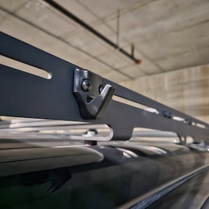 May include: A black roof rack system with a hook-shaped attachment, mounted on a vehicle. The rack has a series of slots and holes, and is designed to carry cargo. The image is taken from a low angle.