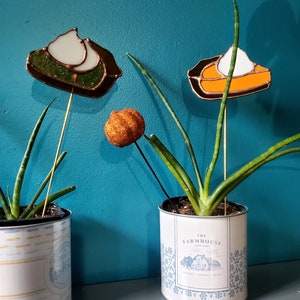 May include: Two potted plants with decorative glass pie toppers. The pie toppers are on metal sticks and are inserted into the soil of the plants. The pie toppers are in the shape of pumpkin pies with whipped cream on top. One pie topper is green and the other is orange. The plants are in white ceramic pots with blue and yellow designs. The pot on the right has the text "The Farmhouse" printed on it.