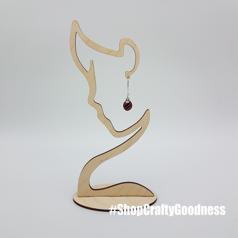 May include: Wooden earring display stand shaped like a woman's profile with a single earring with a red and white striped ball hanging from it. The stand has the text #ShopCraftyGoodness on the base.