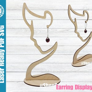 May include: Wooden earring display stand with a silhouette of a woman's head. The stand is designed to hold earrings on the side of the head. The stand is shown with a pair of earrings hanging from it. The stand is labeled "#ShopSmall" on the base.