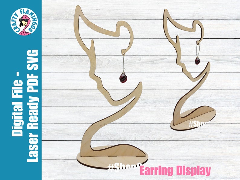 May include: Wooden earring display stand with a silhouette of a woman's head. The stand is designed to hold earrings on the side of the head. The stand is shown with a pair of earrings hanging from it. The stand is labeled "#ShopSmall" on the base.