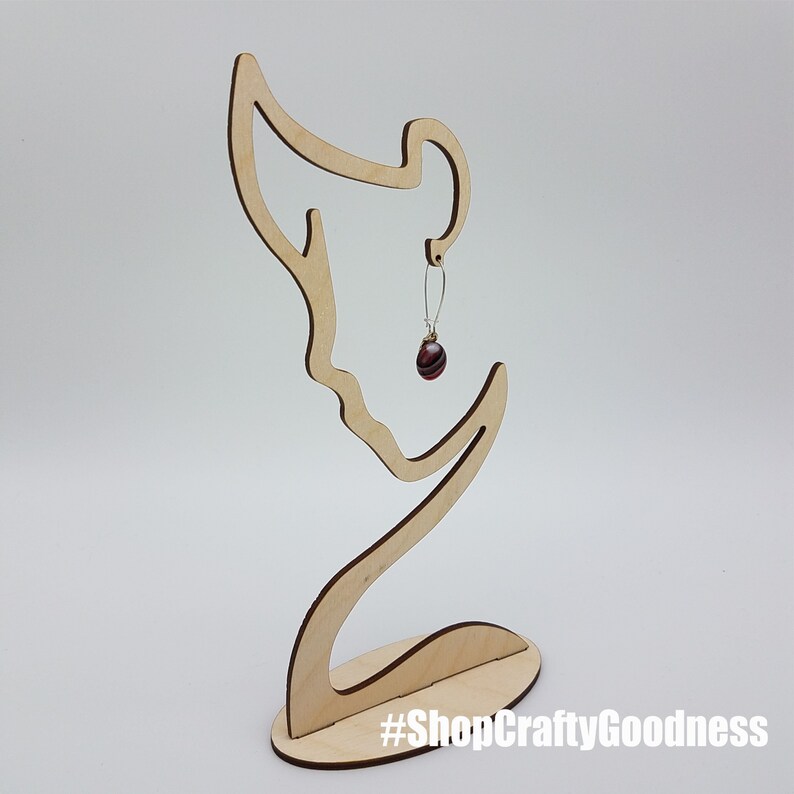 May include: Wooden earring stand shaped like a woman's profile with a single earring hanging from the ear. The earring is a dark red glass bead with silver accents. The stand has a base with the text "#ShopCraftyGoodness".