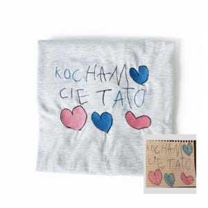 May include: A light grey t-shirt featuring the Polish phrase "KOCHAM CIE TATO" embroidered in blue, with pink and blue hearts. A smaller, handwritten version of the design is visible in the corner.