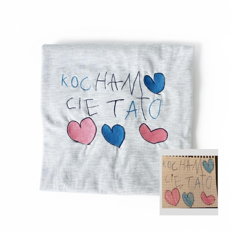 May include: A light grey t-shirt featuring the Polish phrase "KOCHAM CIE TATO" embroidered in blue, with pink and blue hearts. A smaller, handwritten version of the design is visible in the corner.