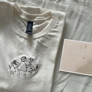 May include: Cream-coloured t-shirt with a crew neck and embroidered black line art of stick figures holding hands. A small drawing of the same figures is next to the shirt. The shirt is folded on a white surface.