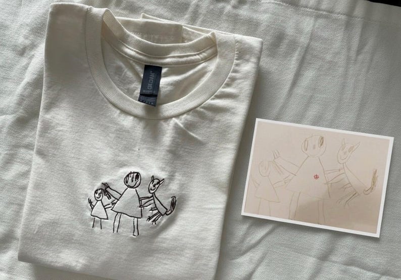 May include: Cream-coloured t-shirt with a crew neck and embroidered black line art of stick figures holding hands. A small drawing of the same figures is next to the shirt. The shirt is folded on a white surface.