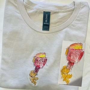 May include: A light grey Gildan Softstyle t-shirt with a crew neck. The shirt has an embroidered design of a colourful, abstract figure. A small drawing of the same figure is next to the shirt.