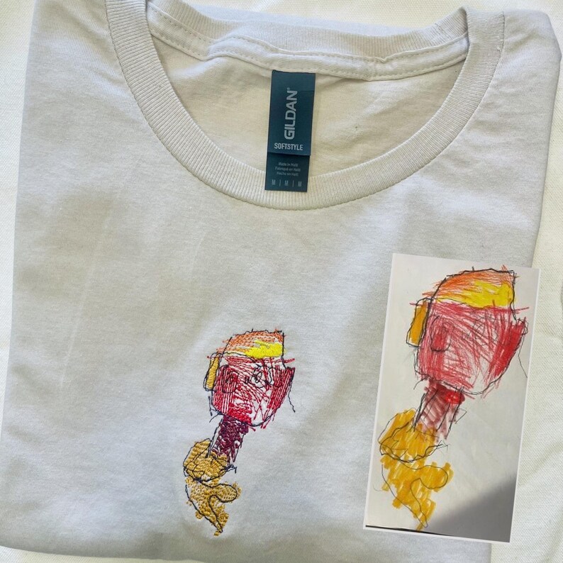 May include: A light grey Gildan Softstyle t-shirt with a crew neck. The shirt has an embroidered design of a colourful, abstract figure. A small drawing of the same figure is next to the shirt.