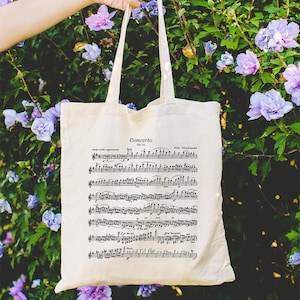 Puede incluir: Una bolsa de lona blanca con notas musicales negras impresas en ella. La bolsa dice "Concerto Op. 64" y "Felix Mendelssohn".