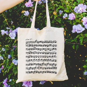 Puede incluir: Una bolsa de lona blanca con notas musicales negras impresas en ella. La bolsa está siendo sostenida por una mano frente a un arbusto de flores moradas.