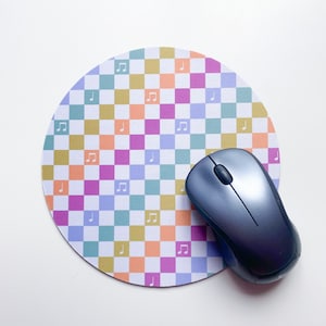 May include: A round mouse mat with a checkered pattern in shades of blue, green, pink, orange, and yellow. The squares have a white background with small music note symbols. A black computer mouse is on top of the mouse mat.