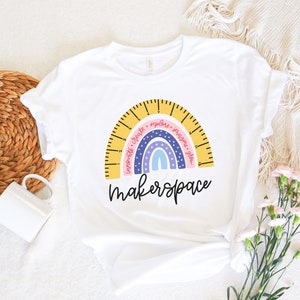 Op de afbeelding: Wit T-shirt met een regenboogontwerp in de vorm van een liniaal. De regenboog bevat de woorden "Innovate, Create, Explore, Imagine, Grow" en het woord "makerspace" in een scriptlettertype.