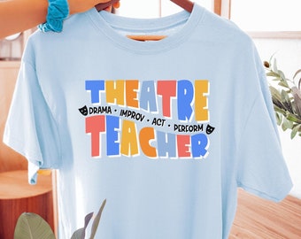 May include: A light blue t-shirt with the words "THEATRE TEACHER" in colorful block letters. The shirt also includes the words "DRAMA, IMPROV, ACT, PERFORM" with small theater mask icons. The shirt is hanging on a wooden hanger.