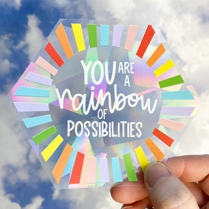 May include: A clear, hexagonal sticker with the text "YOU ARE A rainbow OF POSSIBILITIES" in white script. The sticker has a rainbow-colored design around the edges, with a holographic effect. The background is a cloudy sky.