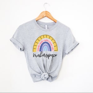 May include: A pale grey t-shirt with a rainbow design. The rainbow has a ruler-like arc with the words "Innovate, Create, Explore, Imagine, Plan." Below the rainbow is the word "makerspace" in a handwritten font. The t-shirt is displayed on a wooden hanger.