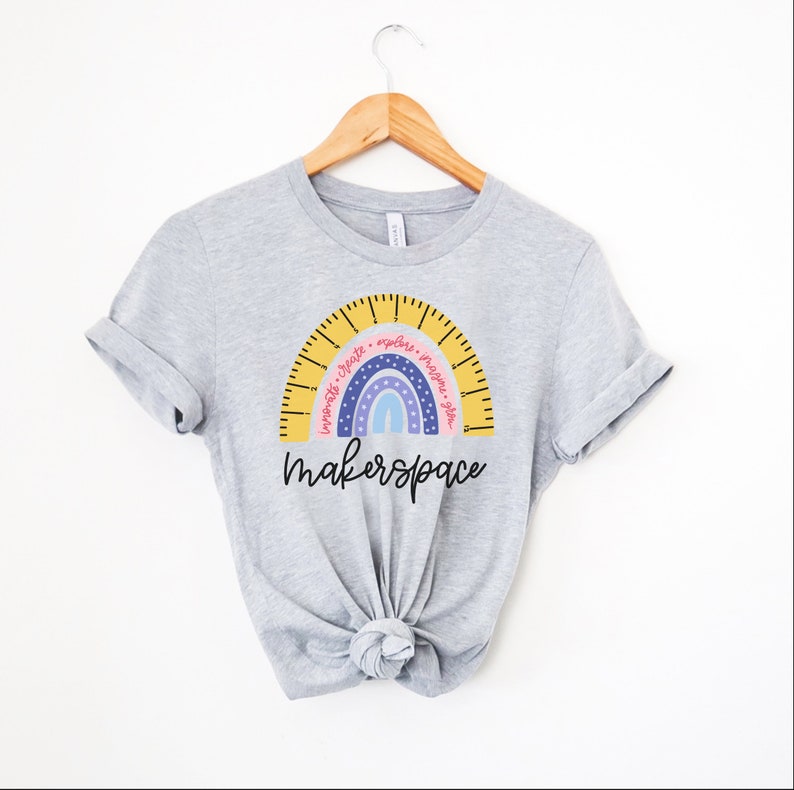 May include: A pale grey t-shirt with a rainbow design. The rainbow has a ruler-like arc with the words "Innovate, Create, Explore, Imagine, Plan." Below the rainbow is the word "makerspace" in a handwritten font. The t-shirt is displayed on a wooden hanger.
