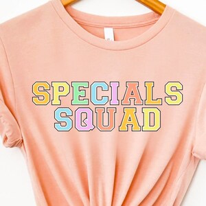 May include: A peach-colored t-shirt with the words "SPECIALS SQUAD" in a colorful, collegiate-style font. The text is outlined in black and filled with pastel colors including yellow, green, pink, and blue. The shirt is hanging on a wooden hanger.