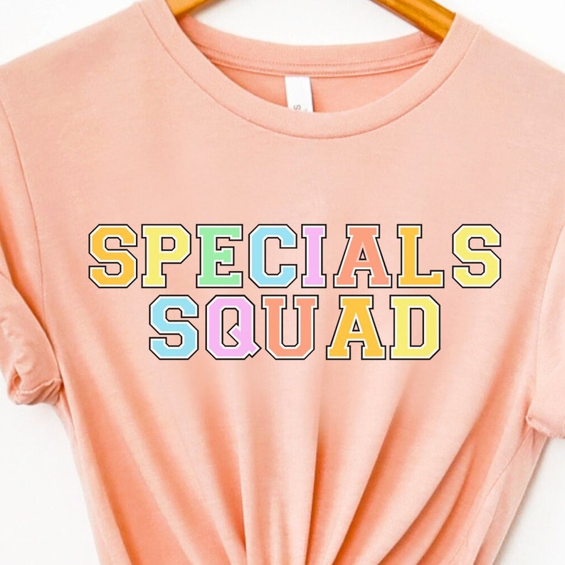 May include: A peach-colored t-shirt with the words "SPECIALS SQUAD" in a colorful, collegiate-style font. The text is outlined in black and filled with pastel colors including yellow, green, pink, and blue. The shirt is hanging on a wooden hanger.
